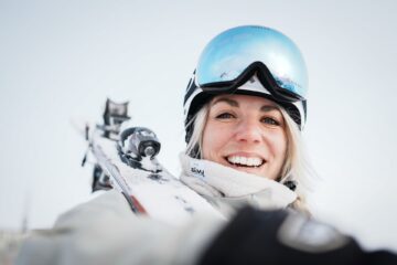 ächelnde Ski-Fahrerin mit Skiern auf dem Rücken und Skibrille auf dem Helm